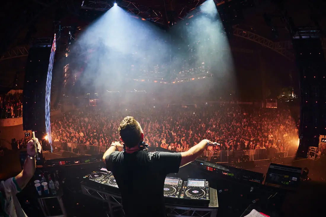 View from behind the DJ, arms out, facing a packed arena